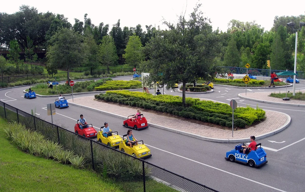 Kinderen rijden in kleine speelgoedauto's op een miniatuurstadsstraat met verkeersborden en rotondes.